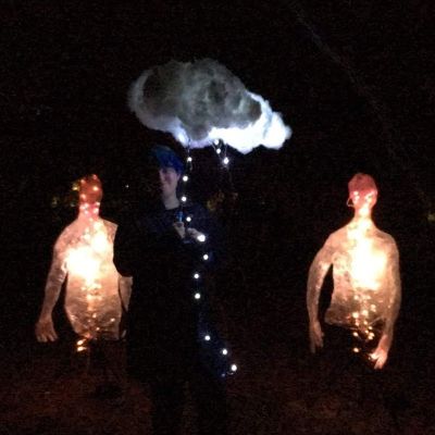 two orange light up humanoid figures on either side of someone holding a lightning storm cloud umbrella art piece